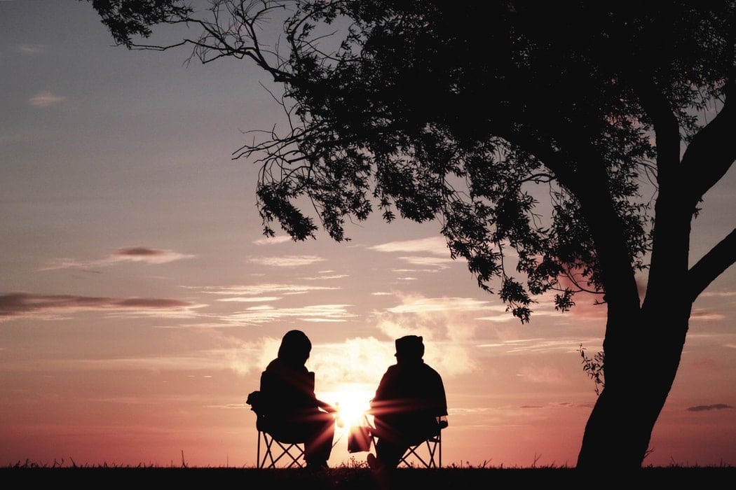Silhouette of two people sitting in camp chairs beneath a tree as the sun sets in front of them.