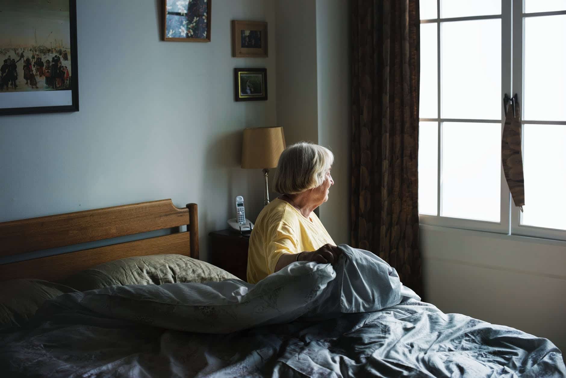 An elderly woman sits in bed, looking out a window. 