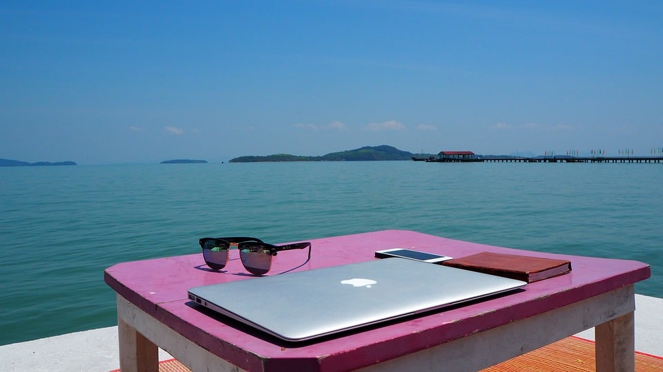 An image of a laptop, cell phone, and sunglasses sitting atop a table, overlooking a body of water. 
